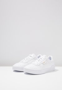 Las zapatillas deportivas blancas presentan una textura suave con detalles perforados, cordones planos y una suela de goma prominente. Acento del logo de la marca en dorado.