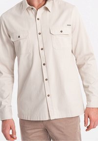 Camisa de color beige claro con botones, dos bolsillos en el pecho, botones marrones y mangas largas; fabricada con tela de algodón texturizada.