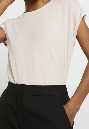 Personne portant un haut beige clair à manches courtes rentré dans un pantalon noir taille haute, avec une main dans la poche.