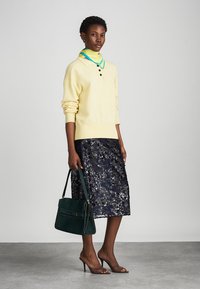 Lys gul strikket trøje med tre knapper, parret med en mørk blomster-mønstret blyantskirt. Accessoriseret med en grøn håndtaske og brune hæle.
