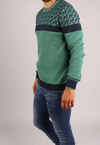 Zielony sweter z przędzy z fakturowanym wzorem w górnej części, ozdobiony granatowymi ściągaczami na rękawach i w pasie, w połączeniu z dopasowanymi dżinsami.