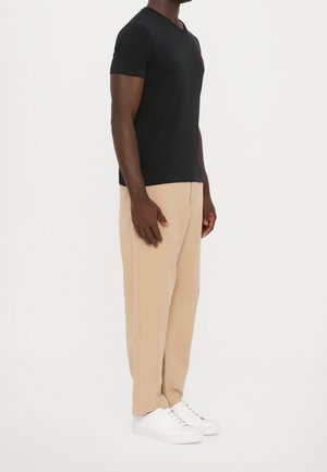 Un homme se tient de profil, vêtu d'un t-shirt noir à manches courtes, d’un pantalon beige et de baskets blanches sur un fond blanc uni.
