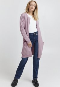 Un long cardigan rose clair avec des poignets et un ourlet côtelés, doté de deux poches avant. Associé à un jean droit bleu foncé et des chaussures noires.
