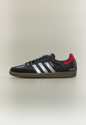 Baskets Adidas Samba noires avec des bandes blanches, un onglet rouge à l’arrière, une semelle en gomme et le logo « Coca-Cola » sur le contrefort du talon.