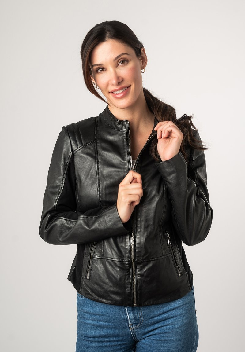 Schwarze Lederjacke mit Kragen, Reißverschluss vorne und seitlichen Taschen; mit gesteppten Details und taillierter Passform. Getragen mit blauen Jeans.