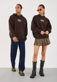 Bruine hoodie met logo, blauwe wijde broek, geruite minirok en zwarte kniehoge laarzen met gespaccenten. Oranje zonnebril op model.