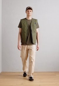 Olive green quilted vest over a dark t-shirt, paired with light beige cargo pants and black sneakers. Simple baseball cap completes the look.