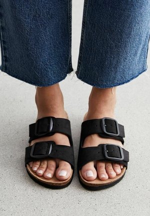 Fødder iført sorte sandaler med dobbelt spænde og lyse neglelakker, stående på en grå gulv, kombineret med blå jeans med frynset kant.