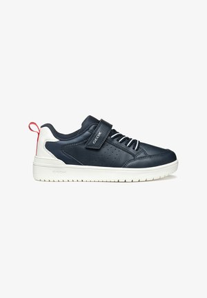 Zapatilla de cuero azul oscuro con suela blanca, con una correa de Velcro, diseño de cordones, perforaciones para transpirabilidad y un acento rojo en el talón.