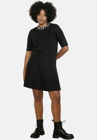 Čierny pletený sveter s krátkymi rukávmi a kvetinovými ozdobami okolo výstrihu, doplnený čiernou sukňou a čiernymi šnurovacími čižmami.