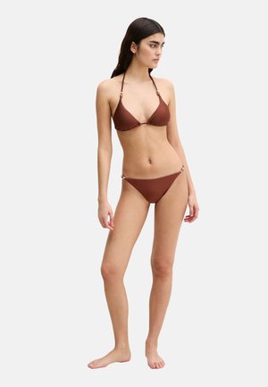 Mujer de pie descalza, vistiendo un bikini marrón de dos piezas con cuello halter y detalles de cuentas a los lados, mirando hacia la derecha sobre un fondo liso.