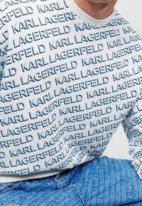 Camicia bianca a maniche lunghe con ripetuto testo "KARL LAGERFELD" in blu. Il tessuto appare liscio con scollatura a girocollo. I pantaloni in denim presentano un motivo di testo simile.