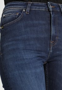 Jeans de mezclilla de color azul oscuro con un ajuste ceñido, que cuentan con una cintura alta, diseño estándar de cinco bolsillos y detalles de costura visibles.