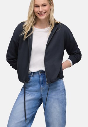 Donna sorridente che indossa una giacca nera con cerniera sopra una maglietta bianca e jeans blu, con le mani dietro la schiena, davanti a uno sfondo semplice.