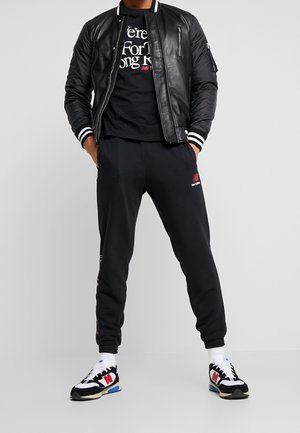 Schwarze Bomberjacke mit weißen gestreiften Bündchen, schwarzes T-Shirt mit Aufschrift, schwarze Jogginghose mit Logo und schwarze Sneaker mit farbigen Akzenten.