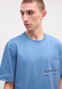 Modrá bavlnená tričko s krátkymi rukávmi a okrúhlym výstrihom. Obsahuje vrecko na ľavej strane hrudníka a vyšívané logo "HUGO" v rôznych odtieňoch modrej.