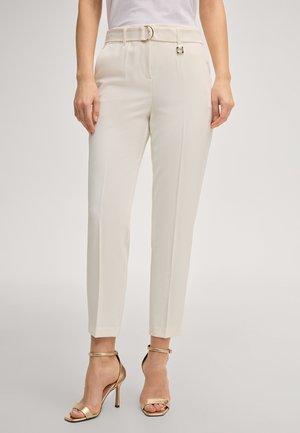 Donna che indossa pantaloni bianchi su misura, corti, con passanti per cintura e sandali dorati con tacco alto, in piedi davanti a uno sfondo neutro.