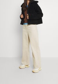 Czarna puszysta kurtka na zamek z kapturem, pod spodem brązowy dziergany sweter, szerokie beżowe spodnie oraz jasne sneakersy z żółtymi akcentami.