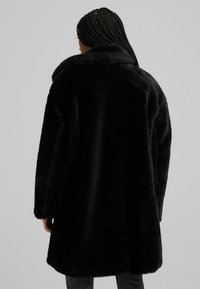 Person with braided hair wearing a long black faux fur coat, facing away against a plain light gray background.