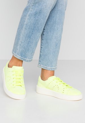 Person iført lyseblå jeans og neongrønne sneakers med hvide såler, stående på en lysegrå gulv mod en ensfarvet grå baggrund.
