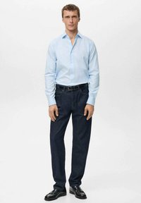 Camisa azul claro, de botones y de mangas largas, combinada con pantalones de mezclilla oscuros. Un cinturón negro y unos zapatos de cuero negro completan el atuendo.