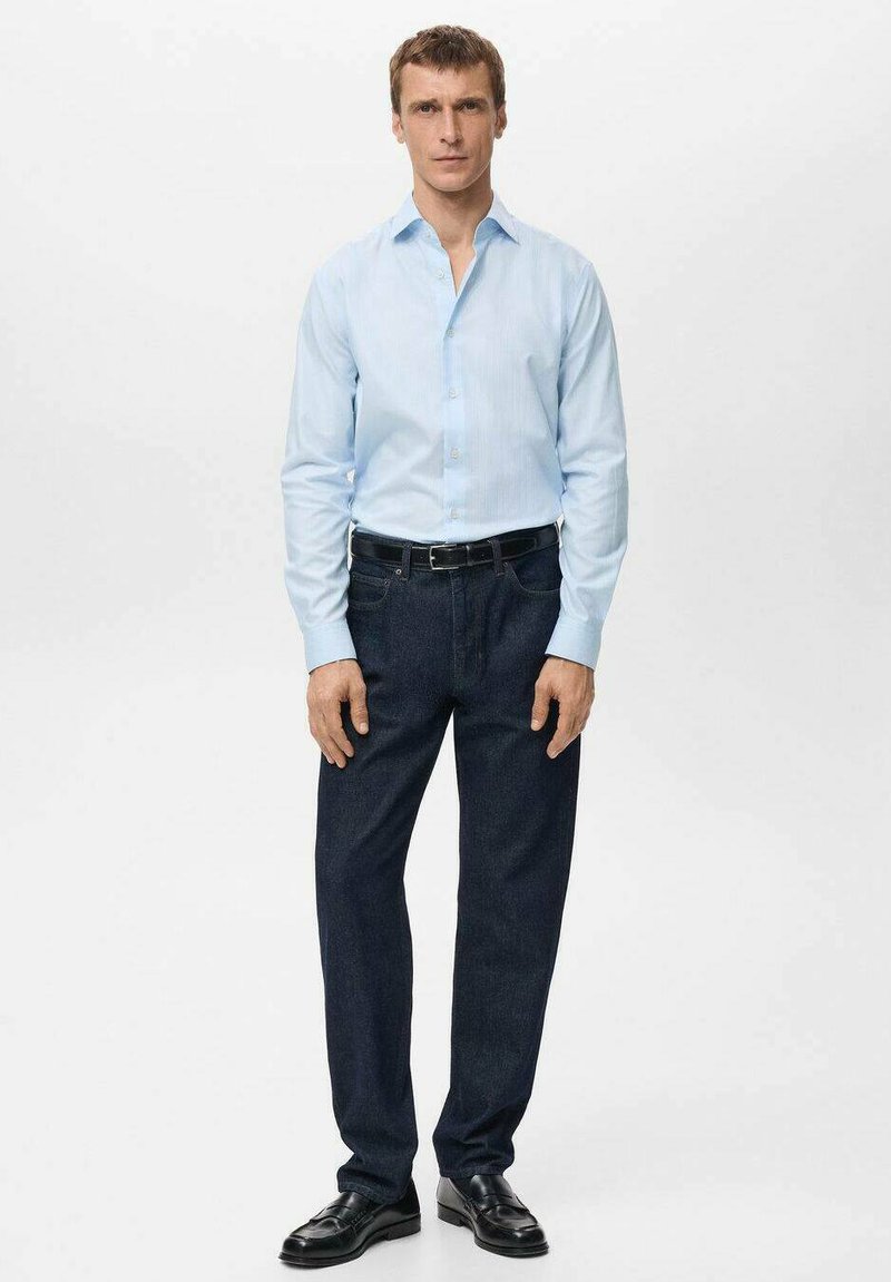 Hellblaues, langärmliges Hemd mit Knöpfen, kombiniert mit dunklen Jeans. Ein schwarzer Gürtel und schwarze Leder Schuhe runden das Outfit ab.