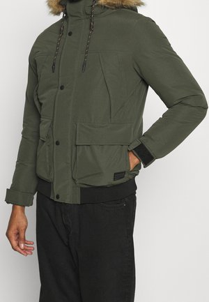 Zielona ocieplana kurtka z odpinanym kapturem wyłożonym futrem, kieszeniami z przodu i ściągaczami przy mankietach. Posiada czarne guziki i kontrastowe przeszycia.