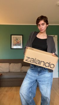Rjava kartonska škatla z velikimi črnimi črkami "zalando" natisnjenimi na njej. Oseba v črnem določenem topu in ohlapnih modrih kavbojkah, ki stoji v notranjosti.