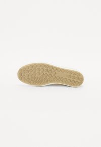 Chaussure à semelle en caoutchouc vue par en dessous avec des motifs de semelle circulaires et ovales pour l’adhérence, sur un fond blanc uni.