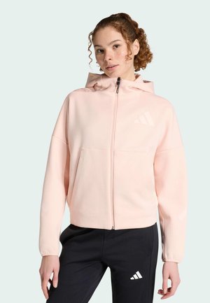 Giovane donna con capelli ricci che indossa una felpa con cappuccio rosa chiaro con zip e pantaloni neri Adidas con logo su sfondo chiaro.