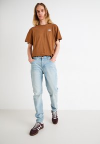 Camiseta de algodón marrón de cuello redondo con un pequeño logo, jeans de mezclilla azul claro y zapatillas de ante morado oscuro con rayas blancas.