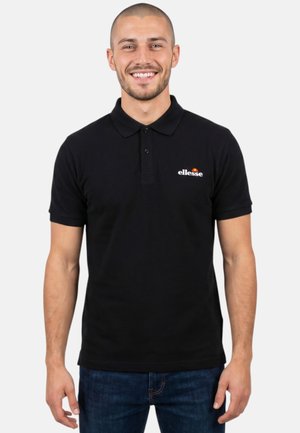 Homme souriant portant un polo ellesse noir et un jean bleu, debout devant un fond clair uni.