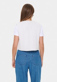 Vit quiltad väst med vertikala ränder, kort design, bärs över en kortärmad vit skjorta, i kombination med högmidjade blå denimbyxor.
