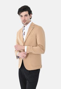 Blazer beige con chiusura a un bottone, revers a listino, una tasca sul petto, tessuto testurizzato, abbinato a una camicia bianca e pantaloni neri.