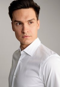 Weißes Hemd mit Kentkragen, glatter Textur und tailliertem Schnitt, sichtbar an einem Mann mit kurzen dunklen Haaren.