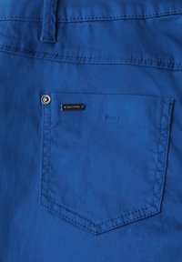 Blauer Stoff mit einer quadratischen Gesäßtasche, die einen silbernen Knopf und ein schwarzes Etikett aufweist. Eine glatte Textur und saubere Nähte sind sichtbar.