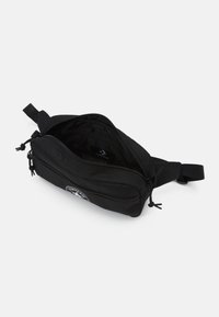 Sac de taille en tissu noir avec un compartiment principal zippé et une poche frontale, doté d'une patch logo rond. Sangle ajustable attachée.