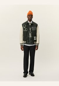 Grøn og creme-farvet varsityjakke med broderede grafikker, stribede manchetter og tal. Parret med en mønstret skjorte og sorte bukser. Orange beanie.
