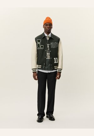 Chaqueta varsity verde y crema con gráficos bordados, puños a rayas y números. Combinada con una camisa de patrón y pantalones negros. Gorro naranja.