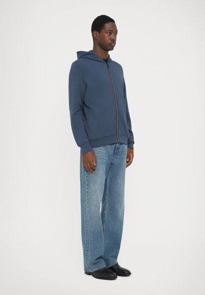 Felpa con zip color navy e cappuccio, con dettagli della zip multicolore, abbinata a jeans a gamba larga blu chiaro e scarpe nere. Sfondo bianco semplice.