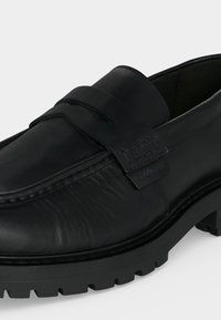 Mocassin en cuir noir avec une semelle épaisse à relief et un détail de bride classique sur le dessus avant.