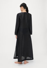 Vestido largo de lino negro con mangas largas, corte recto y textura ligera y translúcida. Cuenta con una abertura alta para facilitar la movilidad y la ventilación.