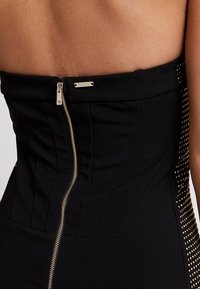 Gros plan d'une robe noire dos nu avec une fermeture éclair centrale dorée et des détails cloutés dorés le long des côtés, portée par une personne.