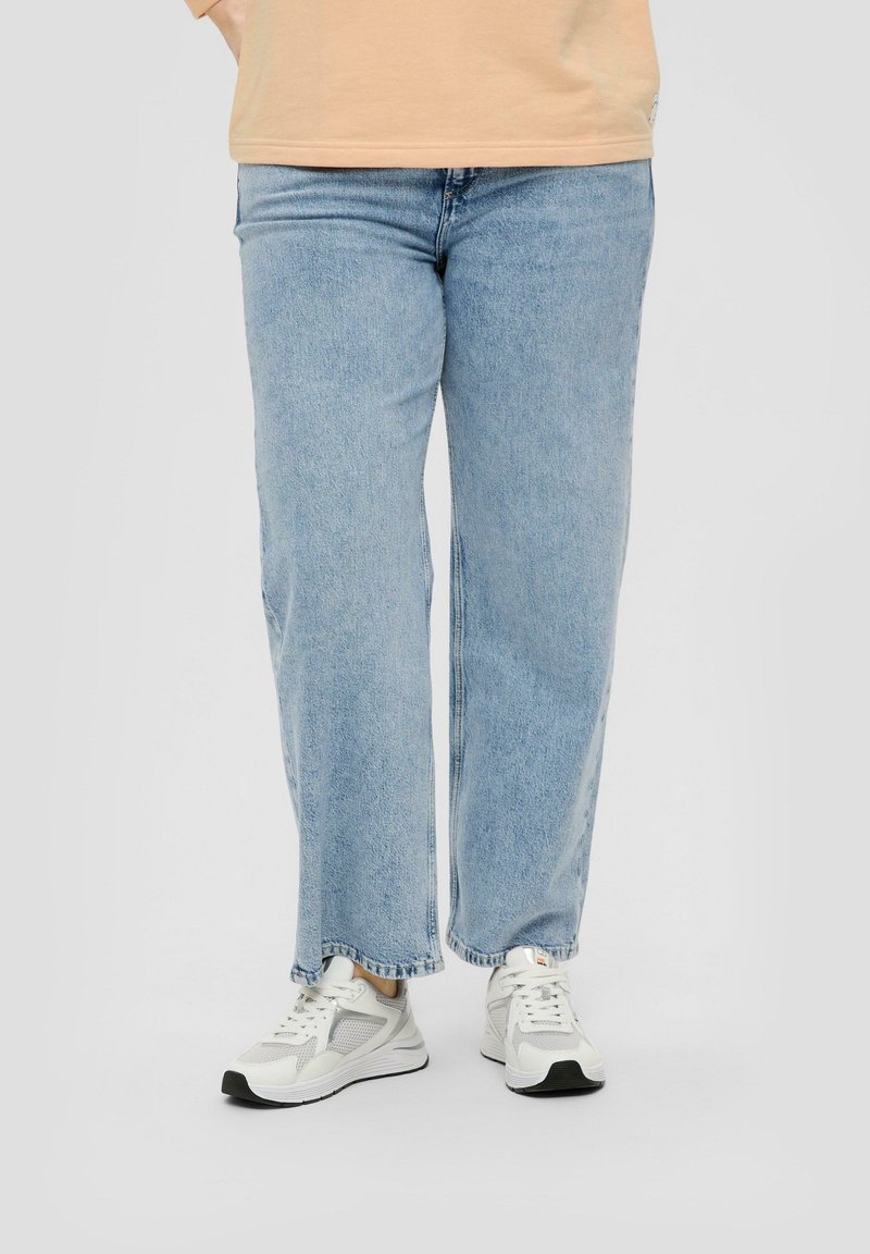 Personne portant un jean droit bleu clair et des baskets blanches, debout devant un fond uni.