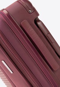 Valise rigide bordeaux avec surface texturée, double zips et nom de marque en relief. Elle présente une poignée en plastique robuste et des accents aux coins.