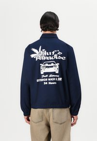 Navy blue jacket with a back graphic featuring white text "Huf's Paradise" and a car design, along with palm tree accents and elastic hem.