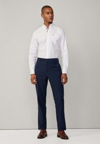 Camisa blanca de botones con un bolsillo en el pecho y un cuello interior a rayas, combinada con pantalones azul marino y zapatos marrones. Textura de tela suave.