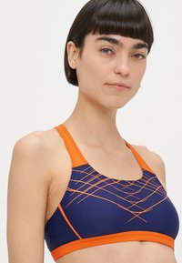 Μπλε σκούρο αθλητικό σουτιέν με πορτοκαλί λεπτομέρειες. Διαθέτει γεωμετρικές γραμμές και σχέδιο με ανοικτή πλάτη τύπου racerback. Κατασκευασμένο από ελαστικό, απορροφητικό υλικό που απομακρύνει την υγρασία.