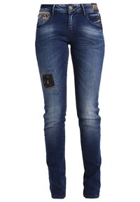 Jean skinny en denim bleu avec un patch déchiré sur la jambe gauche, présentant des coutures élaborées sur les poches avant et une fermeture à bouton.
