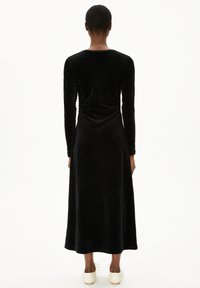Schwarzes Samtkleid mit langen Ärmeln und tailliertem Oberteil, fließend bis zu einem Rock in midi-Länge. Glatte Textur und subtile Raffung.